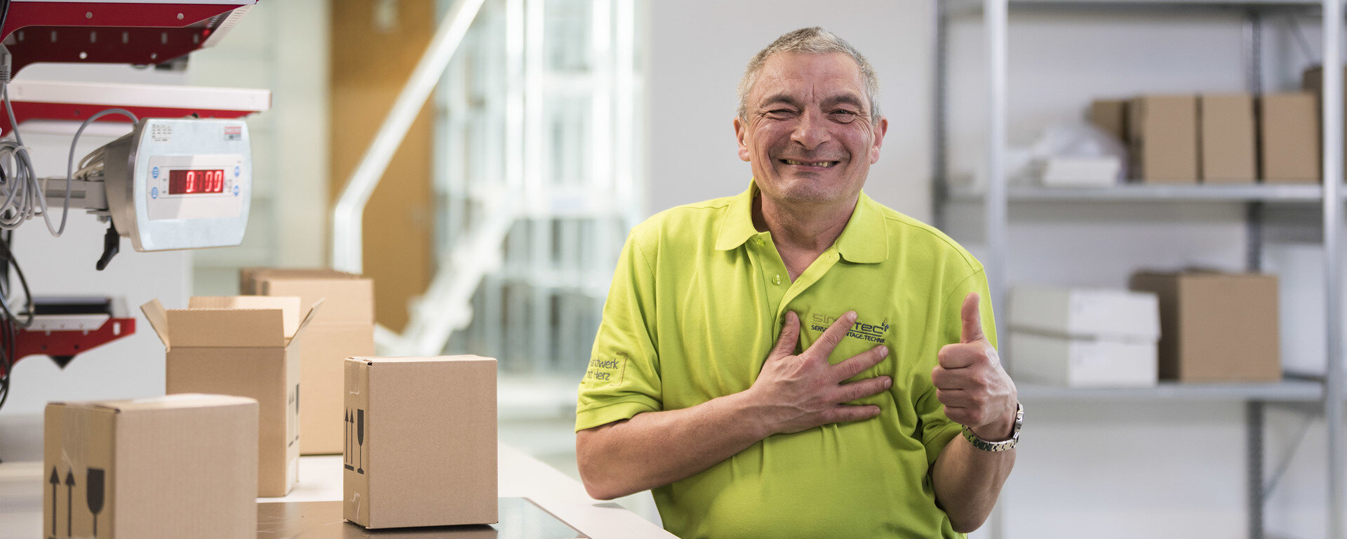 Ein Mann steht in einem Lager mit Kartons neben sich und zeigt einen Daumen hoch