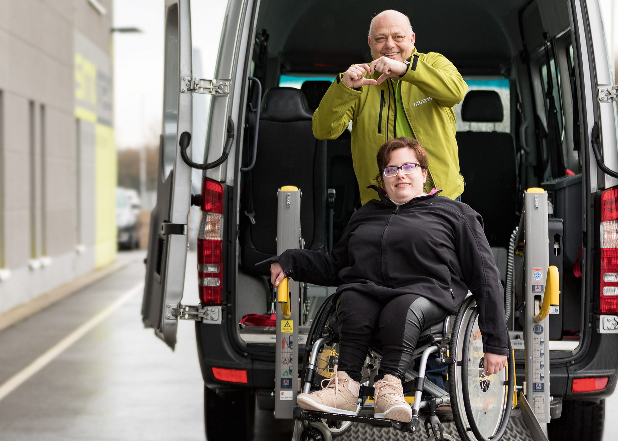 Eine Frau im Rollstuhl auf der Rampe eines Transporters und hinter ihr ein Mann der mit den Händen ein Herz formt