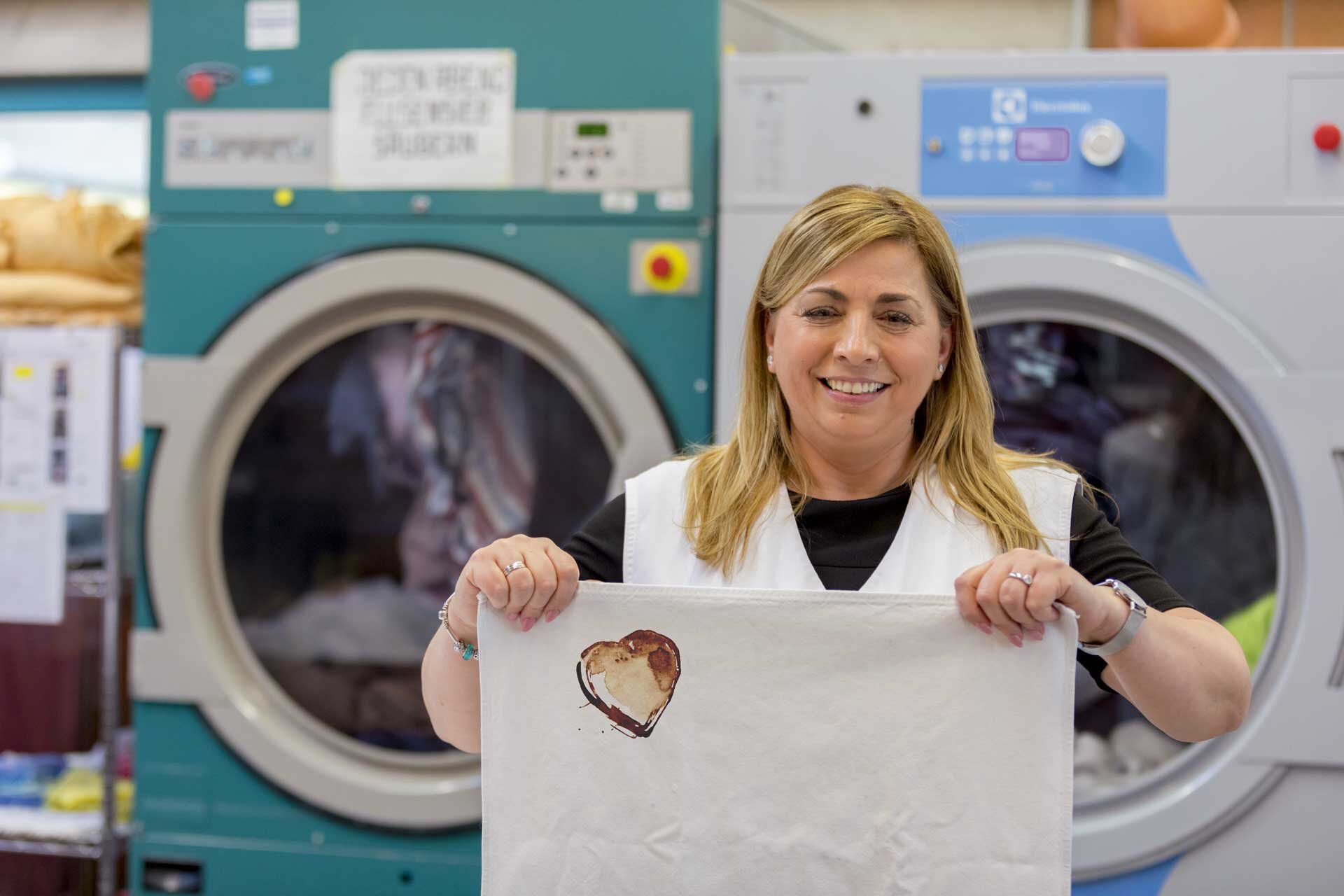 Eine Frau steht vor großen Waschmaschinen und hält ein Tuch mit einem Herzfleck hoch